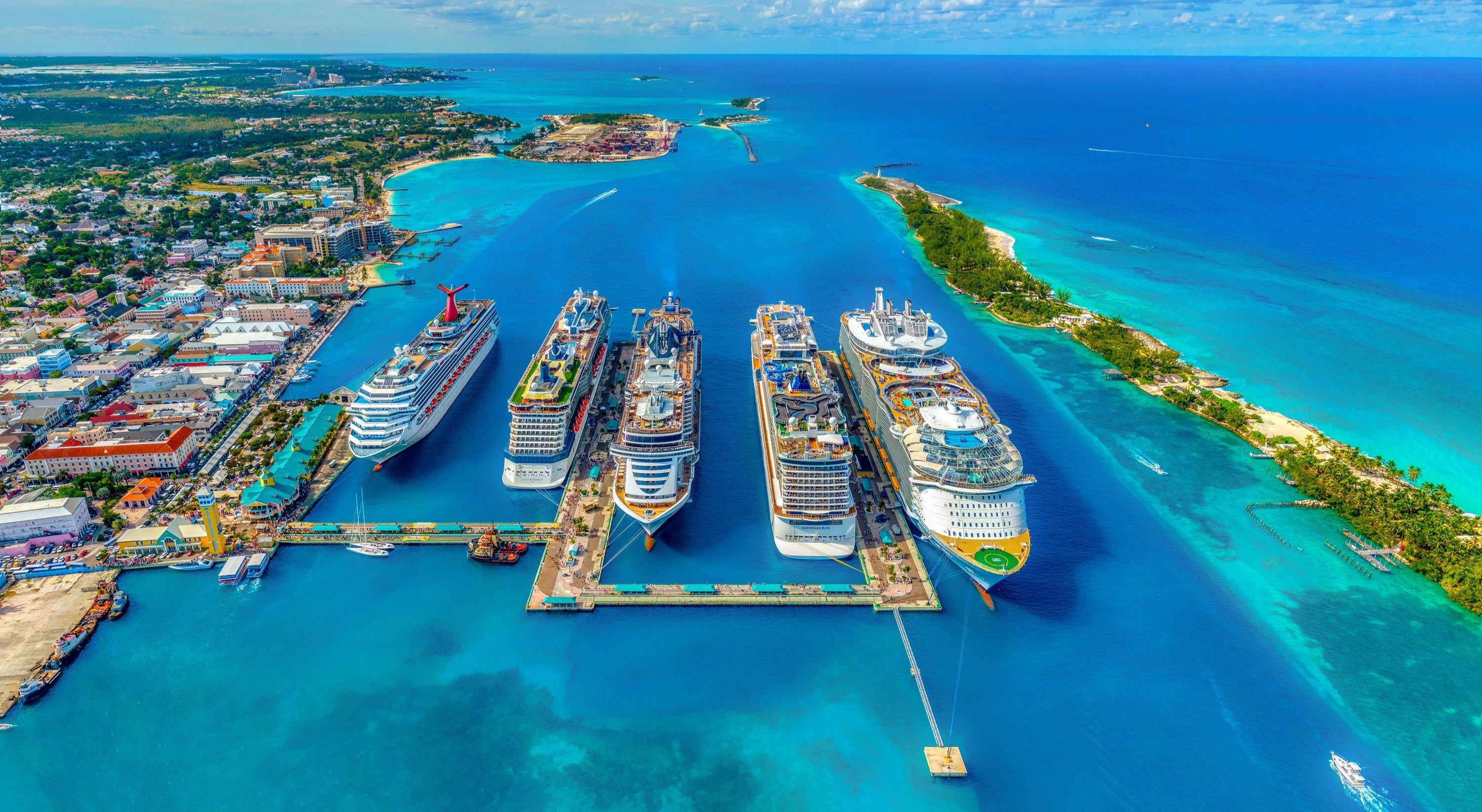 Multiple cruise ships docked at a tropical port with clear blue water and a cityscape in the background.